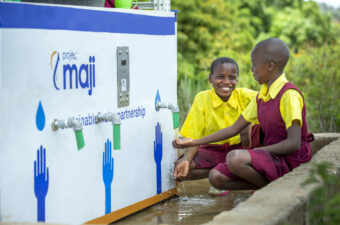 Two African children smiling while getting water from a kiosk