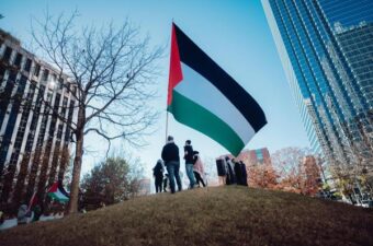 تجمع الناس حول علم فلسطيني كبير على تلة، مع مبانٍ شاهقة وشجرة في الخلفية في يوم صاف.