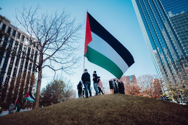 تجمع الناس حول علم فلسطيني كبير على تلة، مع مبانٍ شاهقة وشجرة في الخلفية في يوم صاف.
