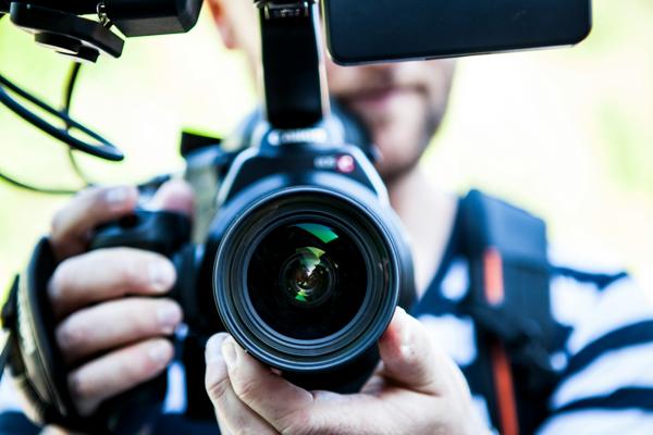 A close-up shot of a person holding a professional video camera, with the lens prominently in focus