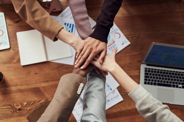 A diverse group of hands stacked together in a show of teamwork and collaboration, with a wooden table, charts, and a laptop in the background